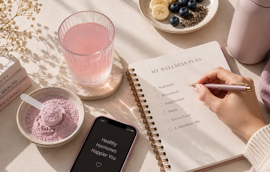 A top-down wellness flat lay featuring a pale pink drink, a bowl of light pink powder with a scoop, an open notebook, a mauve pen, and a hand writing a wellness checklist on a cream stone surface.